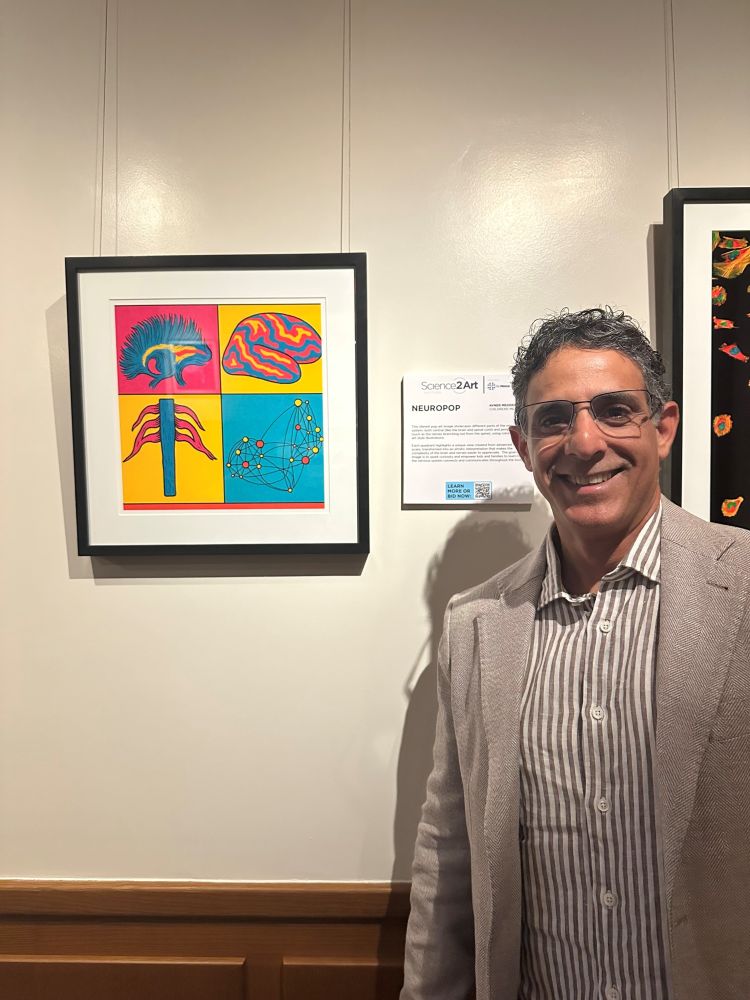Dr. Avner Meoded stands on the right next to his framed scientific art. The art is colorful and has 4 sections.