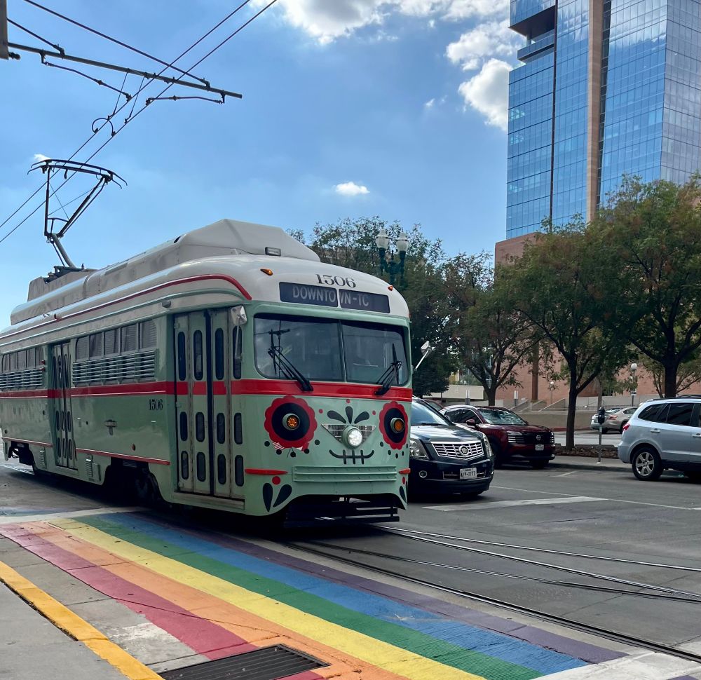 Truly adorably rounded-corner streetcar, the kind with the accordion doors. It’s painted mint green with a nice red stripe for accent. Its front is painted in a Mexican folk art style so it looks like a Día de Los Muertos skull with a cute smile and the headlights are eyes. Bonus: it’s just pulling up to a rainbow crosswalk because the point where the two steeetcar lines cross happens to be I guess near some gay bars. 