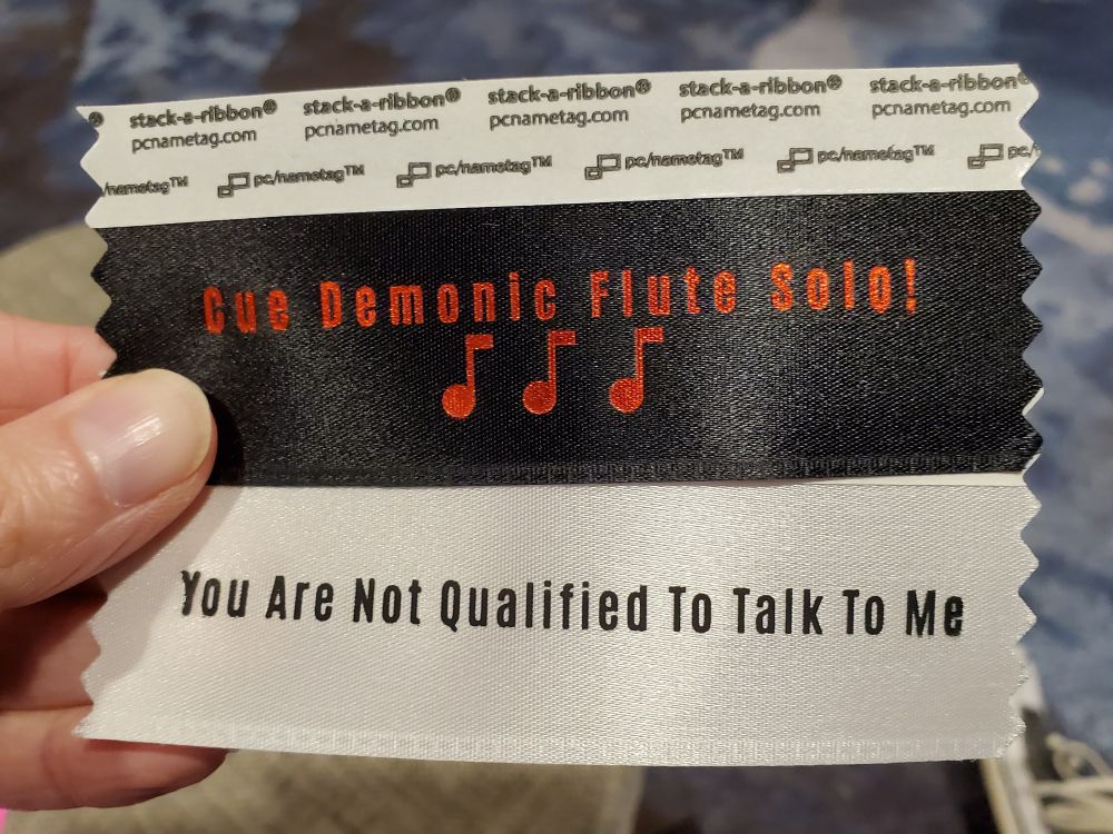 A black badge ribbon with red text reading "Cue Demonic Flute Solo! 🎵" over another badge ribbon that's white with plain black text reading "You Are Not Qualified To Talk To Me"