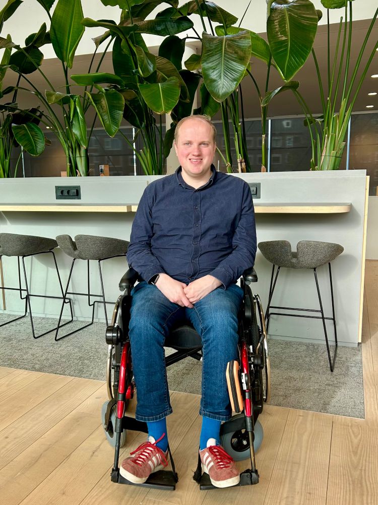 Je ziet mij: witte man met weinig blond haar, donkerblauw overhemd, donkerblauwe spijkerbroek en paarse sokken in rode adidasschoenen met witte strepen. Ik zit in een rolstoel waarvan de stangen rood zijn. Aan de rolstoel hangt naast mijn linkerbeen een oranje tasje. Aan de rechterkant hangt een kastje waarmee ik mijn rolstoel automatisch harder en zachter kan laten rijden.

Ik ben gepositioneerd voor hoge stoelen aan een lange grijze plantenbak met gemonteerde blank houten hangende tafel en zwarte stekkerdozen. In de plantenbak staan hoge planten met zeer grote bladen waarvan ik de naam niet weet