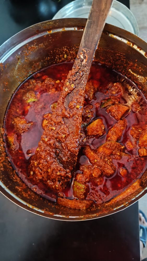 Photo of Avakkai urugai being made. Photo shows a stainless steel vessel with chunks of cut green mangoes being pickled in oil, red chilli powder, mustard powder and salt. It's deep red with some beigish spots.