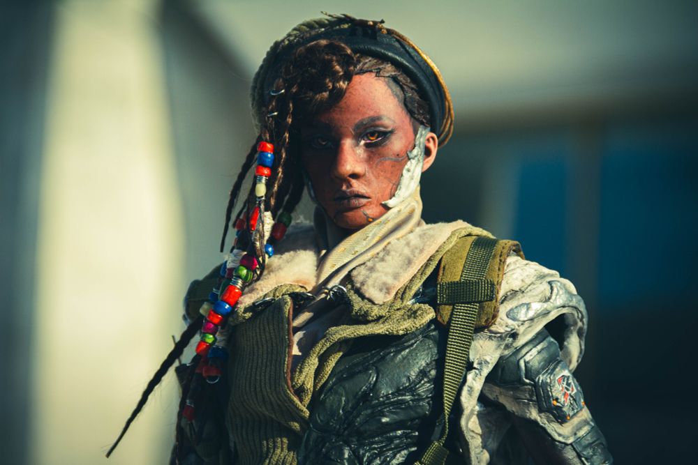 A photo of a BJD doll depicting a black person standing in front of the stairs. They are wearing a military beret and a jacket with sculpted armour parts, and they have beads on their braids.