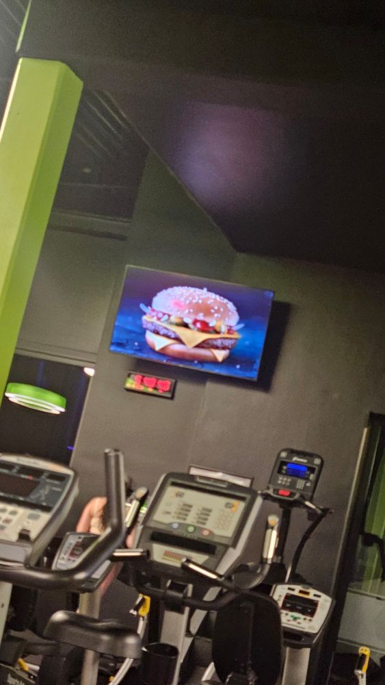 Blick in einen Fitnessrau. An der Wand hängt ein Fernseher. Es ist Werbung für Burger zu sehen.