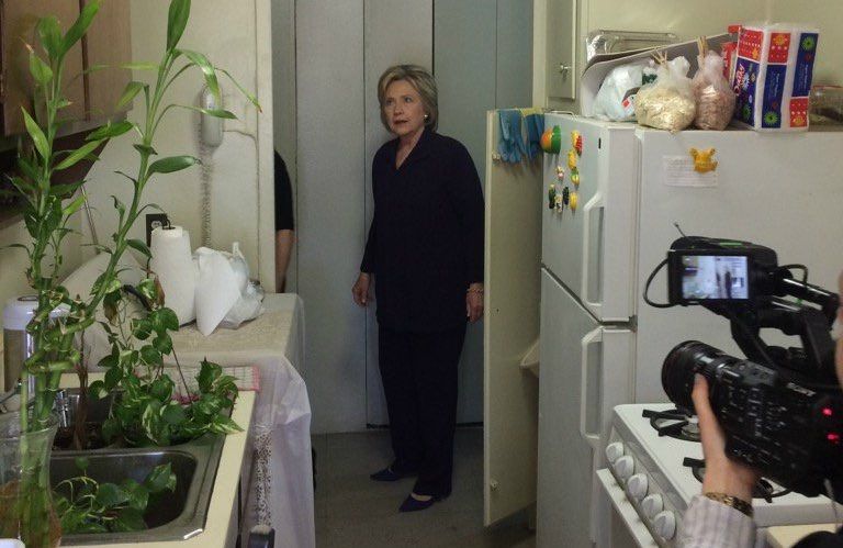 Hillary Clinton standing inside a typical NYC apartment with a tiny kitchen, looking dumbfounded and aghast in front of photographers