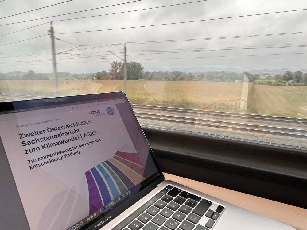 MacBook mit Titelseite des Sachstandsberichts, Blick aus dem Zugfenster auf Bahnstrecke und Felder bei bewölktem Himmel