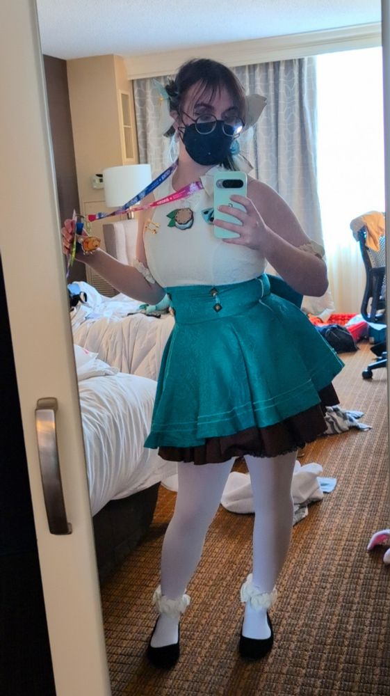im standing in a hotel room, in the same Miku fan art cosplay, with proof of a very full week strewn about the floor behind me. Im holding my badge up in a way that shows off the Oatchi charm hanging off of it