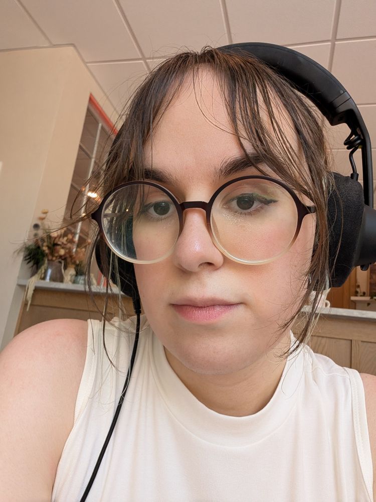 a selfie of me sitting in a coffee shop! it's warmly lit and I'm wearing big over the ear headphones, with a little blush and hair perfectly representative of the humid weather outside