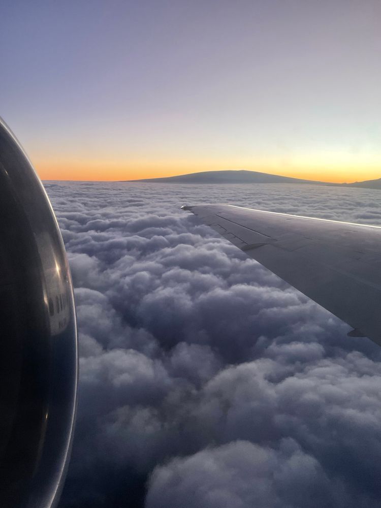 The view from an airplane, depicting a blanket of clouds and a mountain against the sunset 
