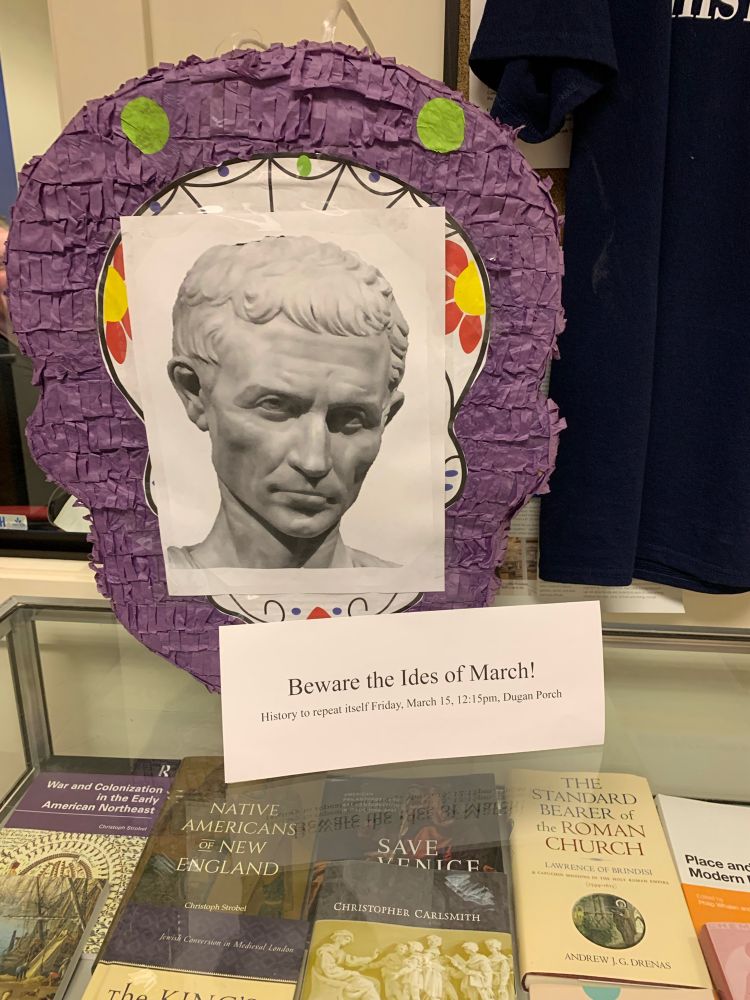 Purple piñata with the image of a bust of Julius Caesar sits on a table. In front of it, a sign reads “Beware the Idea of March! History to repeat itself, Friday, March 15th”