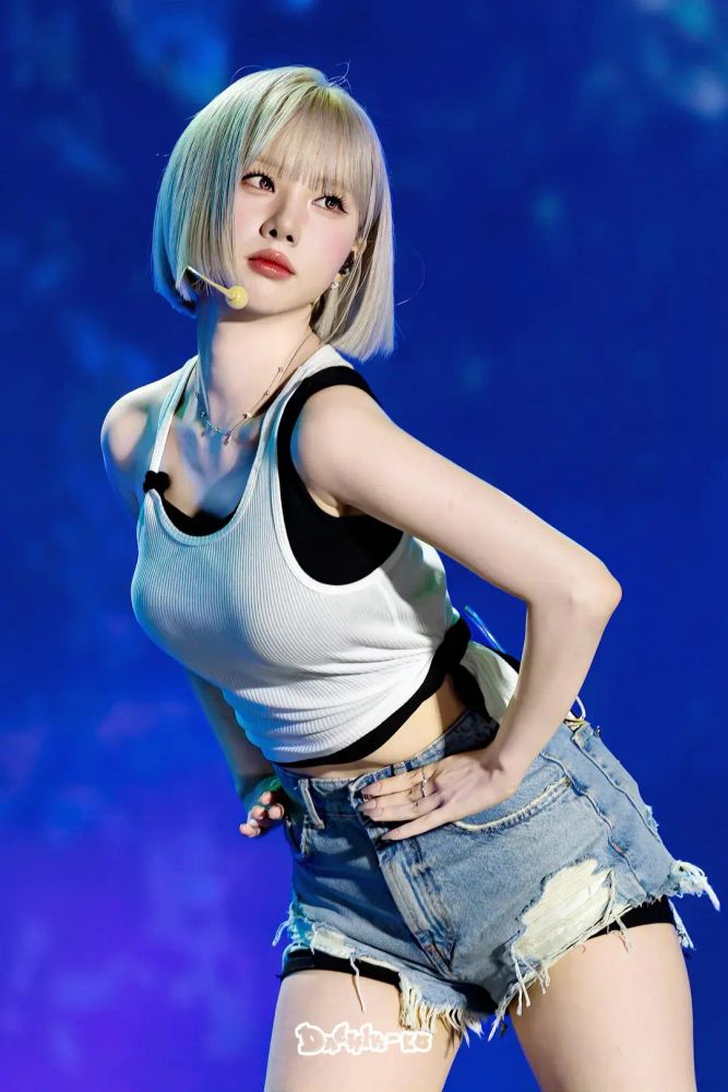 Kpop idol Eunha  in a three quarter length still from  her group VIVIZ in performance. She wears frayed denim shorts and a sleeveless white crop top beneath which a black crop top is visible. Around her neck is a silver necklace. Her blonde  hair is styled in a bob with bangs. She is leaning forward, placing her hand near her hips, and putting her weight on her right leg as she turns and inclines her  head back a little to look over her left shoulder.