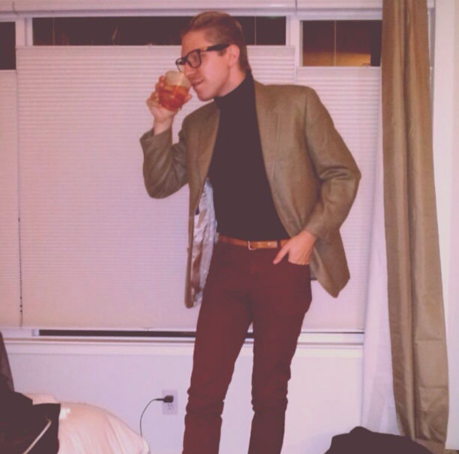 a young man in slim oxblood pants with a light brown skinny belt, a charcoal turtleneck, a tweed blazer, and fake thick black glasses drinking an old fashioned 