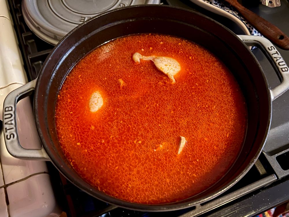 a dutch oven with a vibrant, menacing red stock with glassy oil on top and semi-poached chicken thigh meat sticking out in spots