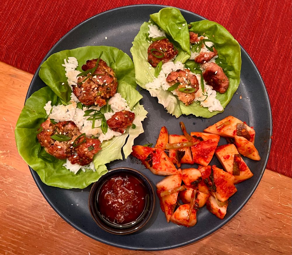 two large butter lettuce leaves topped with sticky rice, broken up meatballs, scallions, and sesame seeds on a plate with a dish of a red paste in salted oil and large chopped pieces of vibrant red radish