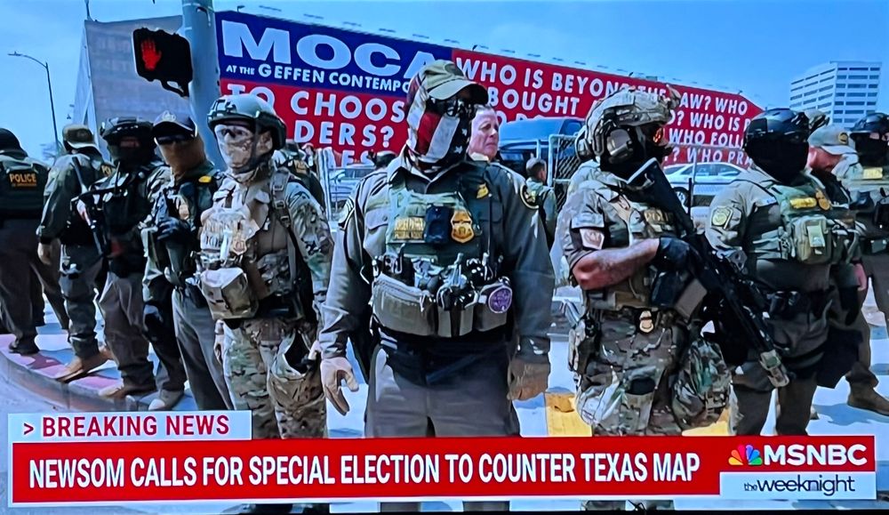 armed and masked cbp agents in full gear and masks, one dead center in an american flag mask , arrayed along an urban street corner in front of a red white and blue text mural. below is a chyron: BREAKING NEWS NEWSOM CALLS FOR SPECIAL ELECTION TO COUNTER TEXAS MAP MSNBC