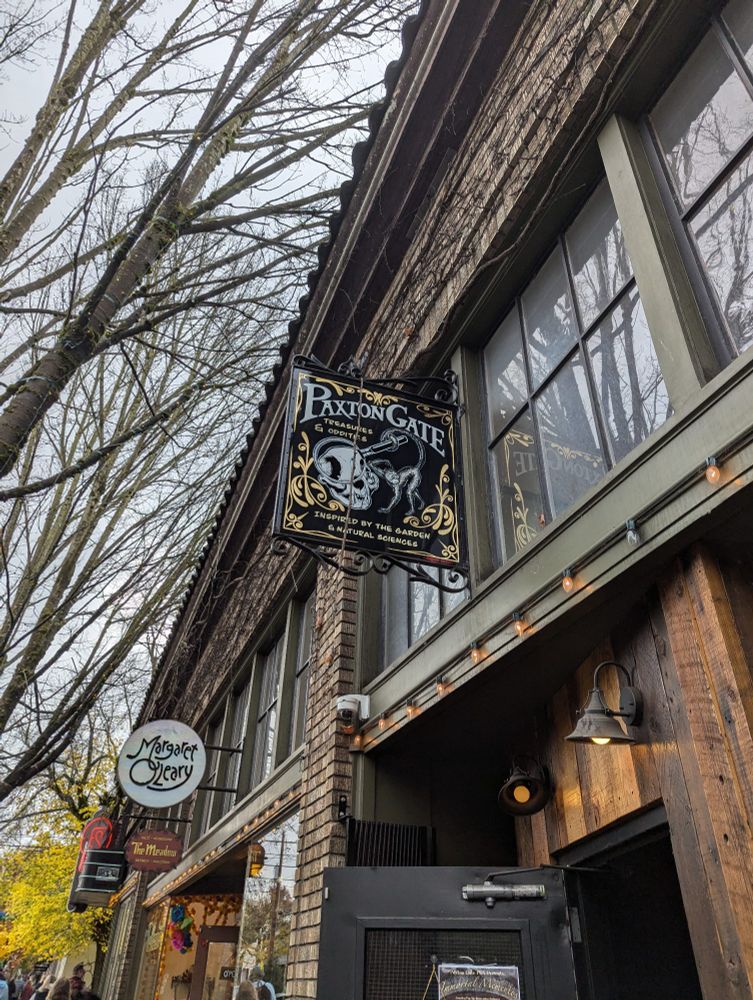A brick front building rises on the right, stretching down to the left as it goes into the background. On the left the branches of naked trees rise into a white sky. 
In the lower right, a door is open on the building, the interior dark against the brown wood facade of the shop. Above the door a sign reads Paxton Gate in white gothic lettering. The edges of the sign are outlined with symmetrical golden filigrees. Below the title in a smaller script reads treasures and oddities. Below that a cat skeleton holds a magnifying glass with its tail over its face making that portion of the skull larger. At the bottom of the sign in small script it reads inspired by the garden and natural sciences. 