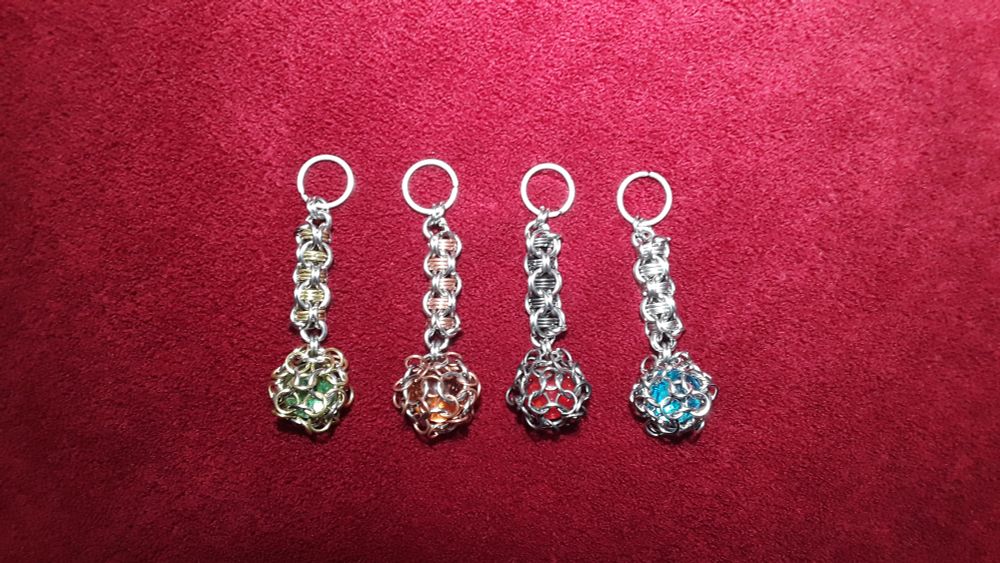 Four chainmail keychains with captive marbles - green, orange, red, and blue - symbolizing the seasons of a year, on a red velvet background.