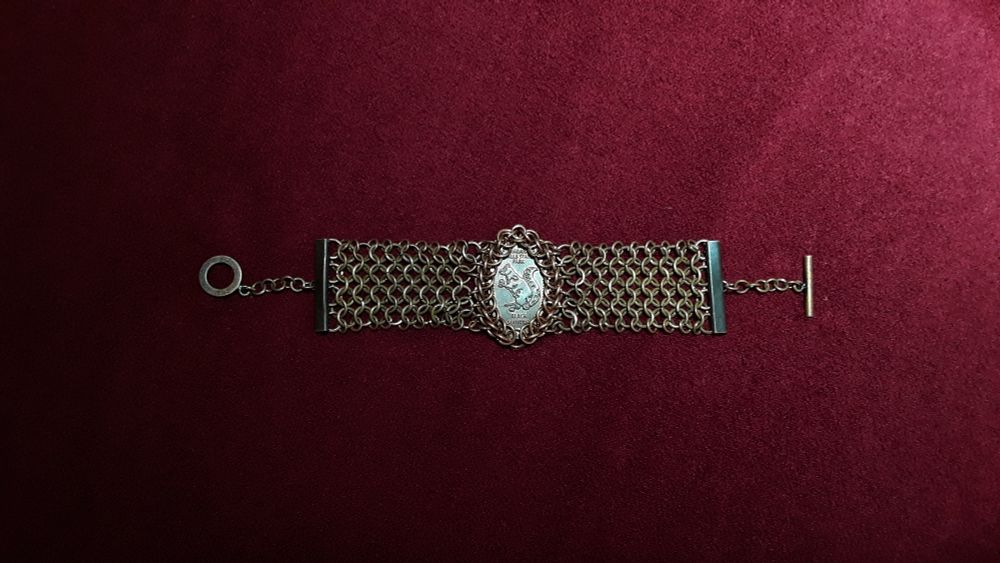 A copper chainmail bracelet on a red velvet background. Design is similar to a watch strap, with a band of European 4-in-1 weave and a 3/4 Persian weave encircling a re-minted penny token, displaying a black squirrel from the Niagara State Park.