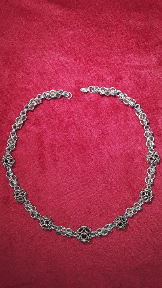 A chainmail necklace with copper accents and black glass marble beads, on a red velvet background.