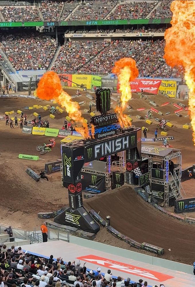 Seth Hammaker crossing the finish line in the 250cc dirt bike main event jumping over a giant dirt mound while flames explode around the finish line banner in a giant sports stadium 