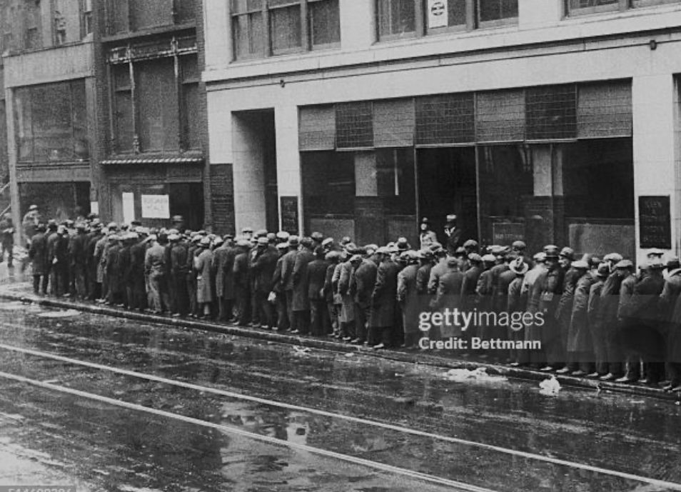 Black and white photo of people standing on a sidewalk waiting in a bread line during the Great Depression. 