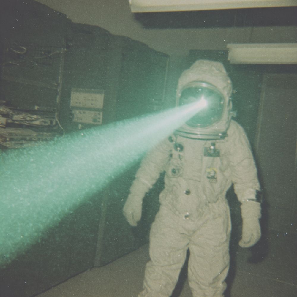 The image depicts a person in a full astronaut suit standing in a dimly lit room, likely an indoor setting. The room features equipment and storage units along the walls. The most striking feature is a bright, greenish light beam emanating from the helmet's visor, casting a glow across the room. The suit is bulky and textured, including various connectors and patches. Overhead, fluorescent lights can be seen on the ceiling. The room’s subdued lighting creates a mysterious atmosphere, while the light beam from the helmet adds an element of intrigue.
