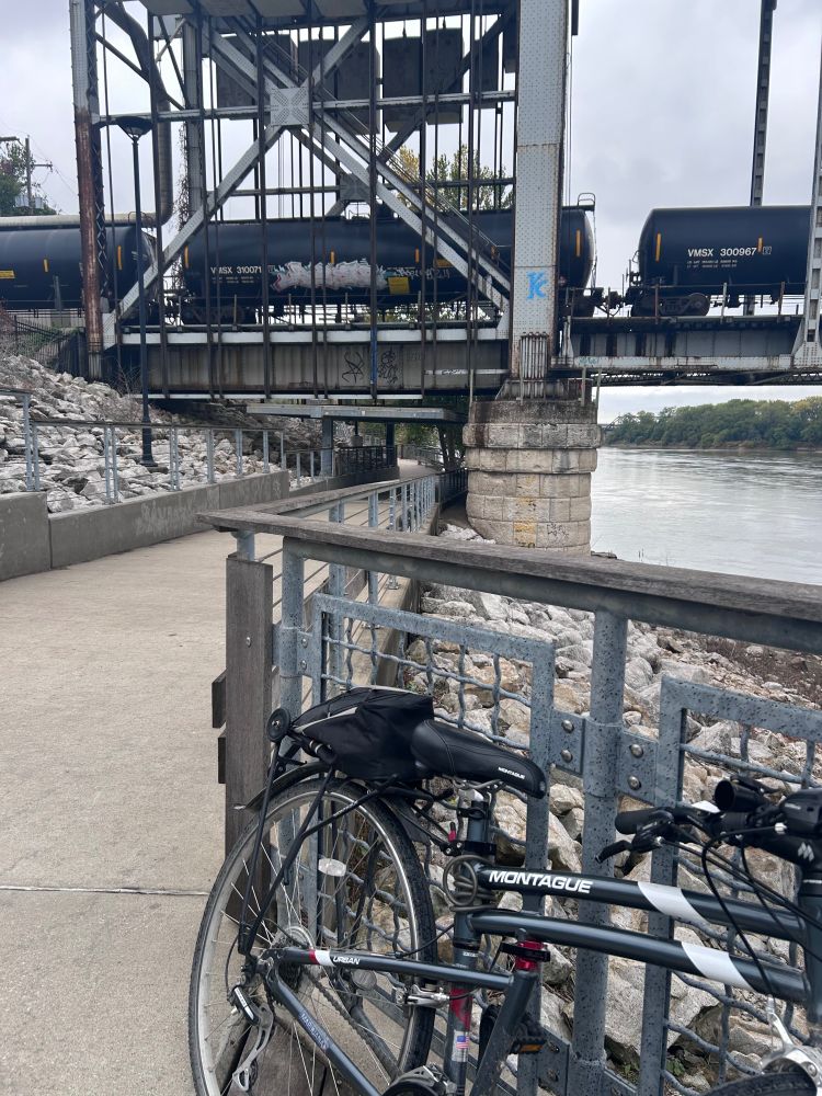 a folding montague bike leaning against a metal fence while several DOT-111 rail cars transport thousands of gallons of ethanol across the Missouri River
