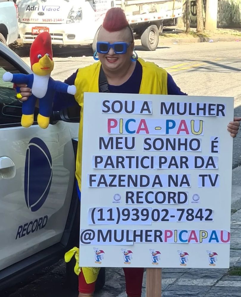 Mulher pica pai na frente do carro da Record segurando um pica pai de pelúcia em uma mão e na outra um cartaz escrito "Sou a mulher Pica-Pau. Meu sonho é participar da Fazenda na TV Record (11) 9 39027842 @mulherpicapau"