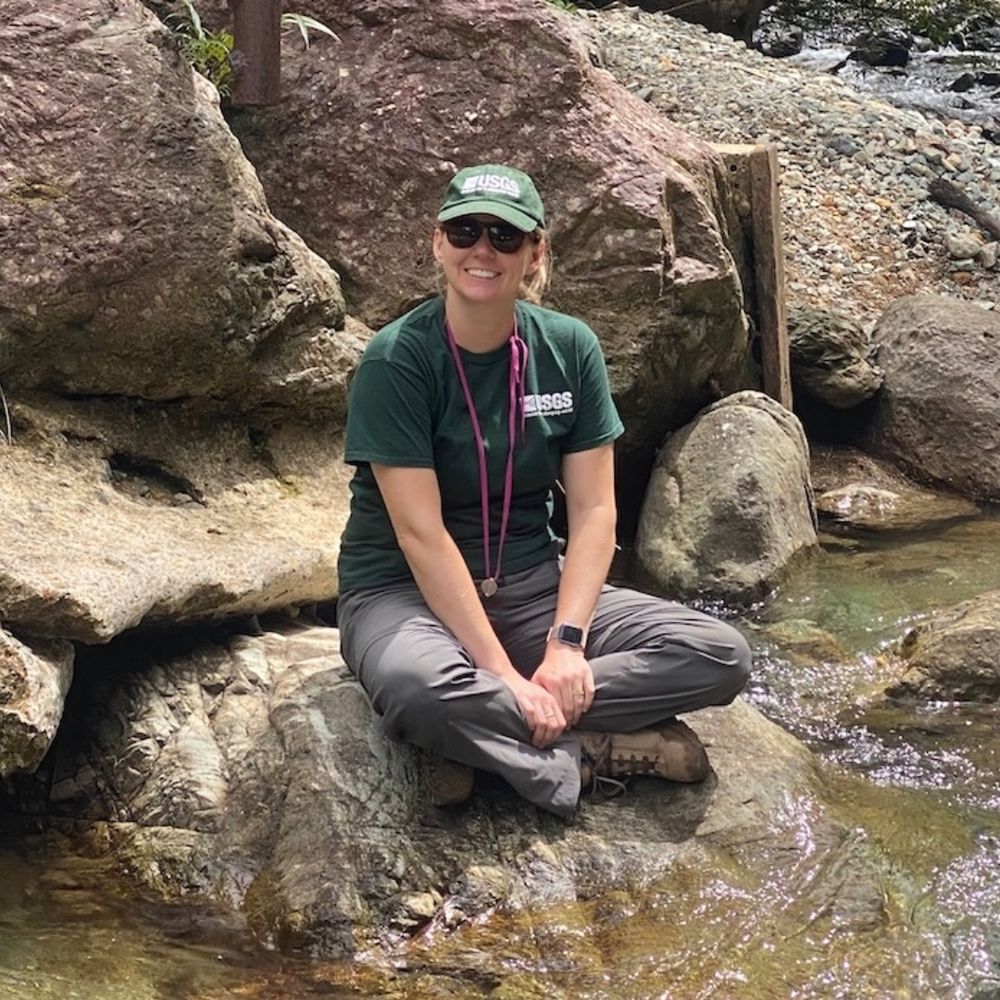 A woman sits on a rock by a river wearing green USGS hat and shirt.