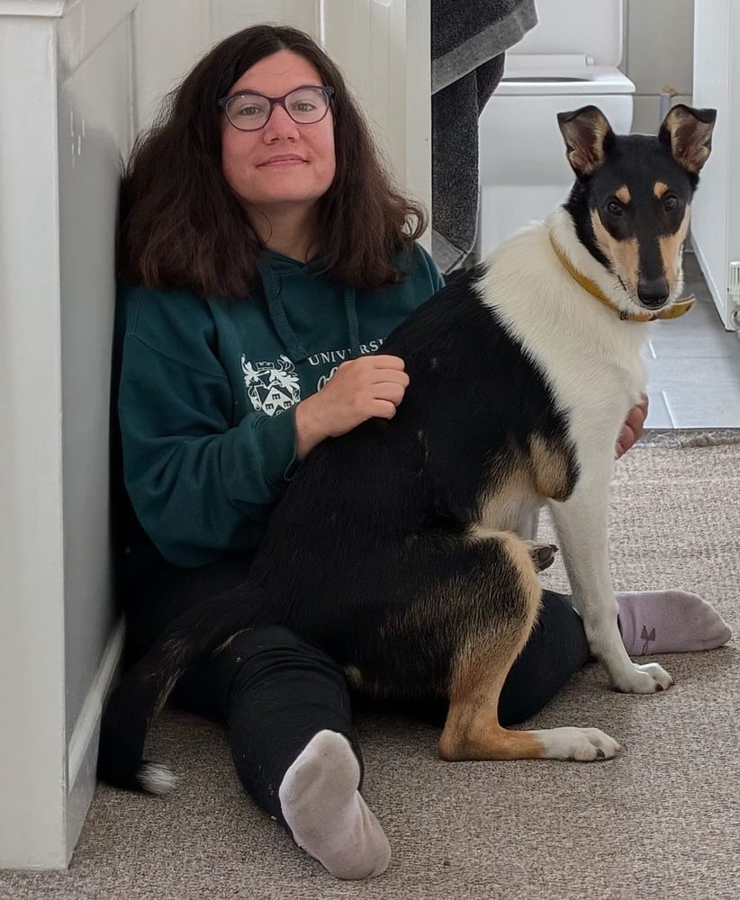 Me (a mid-thirties short, brown haired woman wearing dark glasses, a green hoodie and black work-from-home pants, with a wide nose and a big smile, looking straight at the camera) sitting on my hallway floor with my dog, Crick(et), a 7-month old tricolour smooth collie. Crick(et), who is as tall as me sitting down, has decided to sit on my lap. He does not fit on my lap. He’s taken to doing this anytime I pet him on the floor.