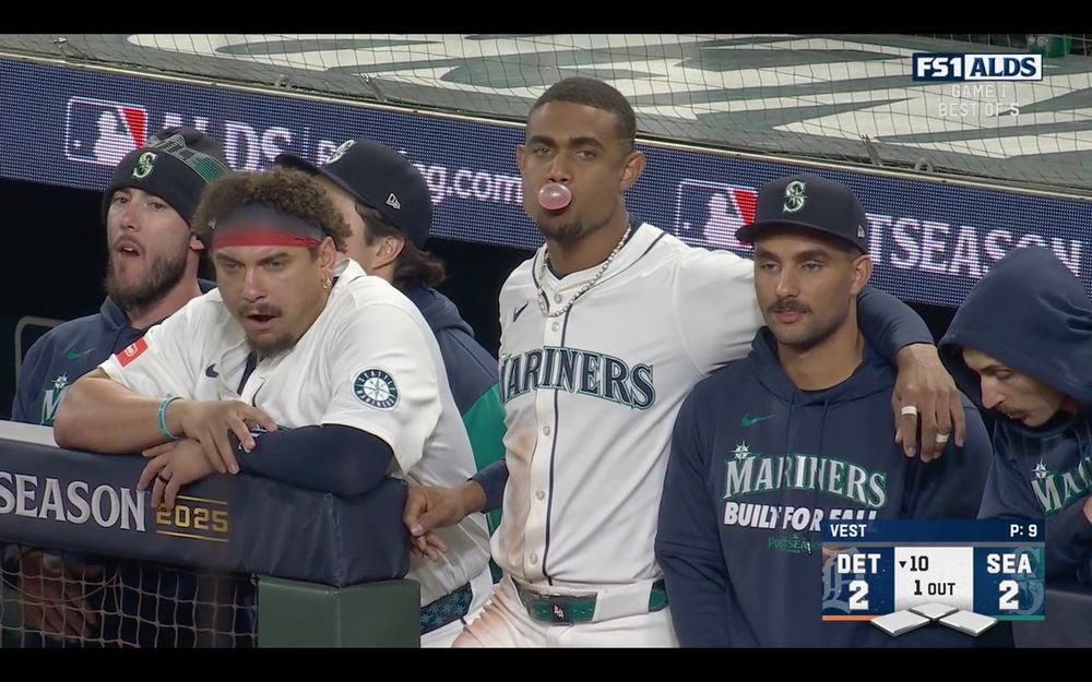 Screenshot of Mastrobuoni, Naylor, Julio and Ford in the Mariners dugout. Miles and Josh appear to be working on some sunflower seeds. Julio is blowing a bubble and has his arm around Harry’s shoulder.