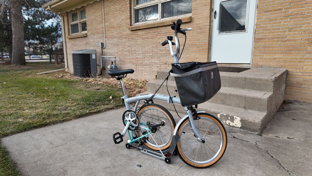 A grey-and-seafoam Brompton folding bike, half set up because there's no kickstand on this thing... yet. It's on the back porch of my office.