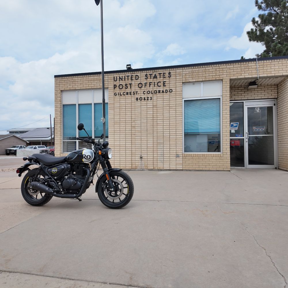 A black and silver Royal Enfield Hunter 350, in front of a rural post office. A tiny, tan brick building, two big windows out front with a blue tint. The sky above is overcast. 



CORN BRO. 