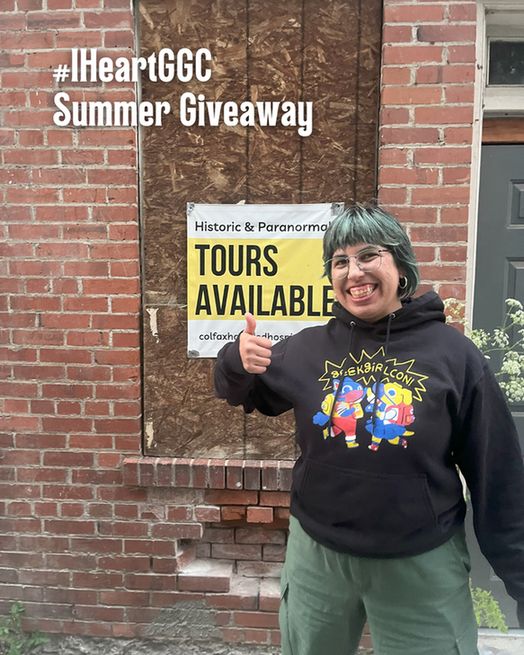 #IHeartGGC Summer Giveaway: Marina, in GeekGirlCon hoodie and a big smile, gives a thumbs up in front of a brick wall and a sign for historic and paranormal tours.