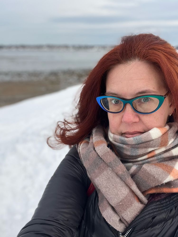 A red-haired woman wearing green glasses, a plaid scarf and a black coat standing outside with snow and the ocean in the background.