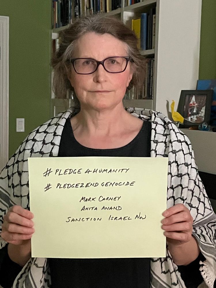 A woman in a kuffiyeh hods a sign reading '#pledge4humanity #pledge2endgenocide - Mark Carney Anita Anand - Sanction Israel Now'