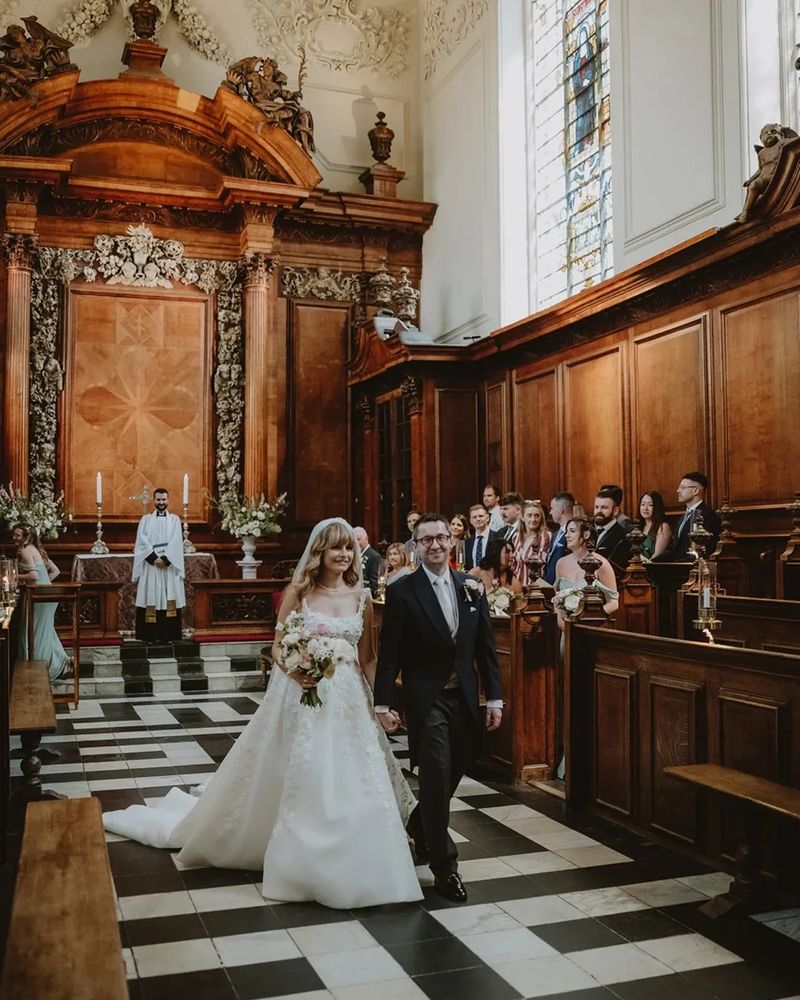 Bride and groom Laura and Conor walking hand in hand down the aisle of Trinity College chapel, surrounded by guests and ornate wooden panelling. A priest stands smiling at the altar behind them beneath decorative carvings and a stained-glass window.