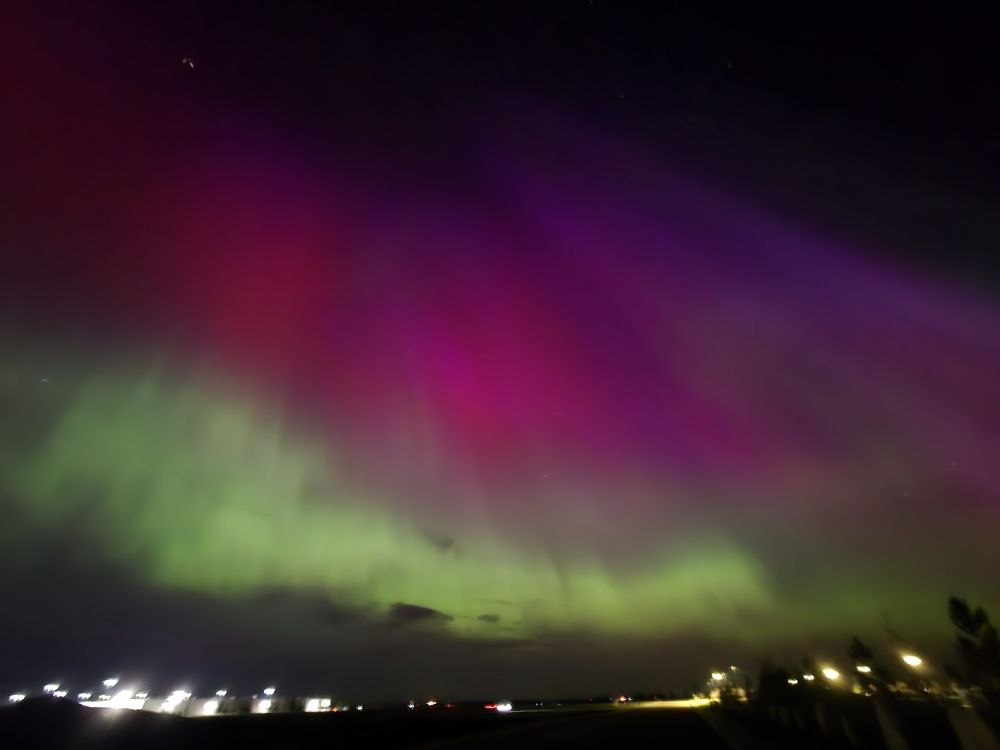 A photo of aurora borealis. Deeply saturated purples and pinks streak across the sky, with the bright lights of a town in the lower left corner of the image.