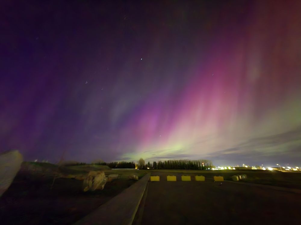 A long-exposure image of aurora borealis that mostly shows the deep purples. The lights of a town are visible in the lower right corner of the image. Some pinks and greens are also visible on the right side.