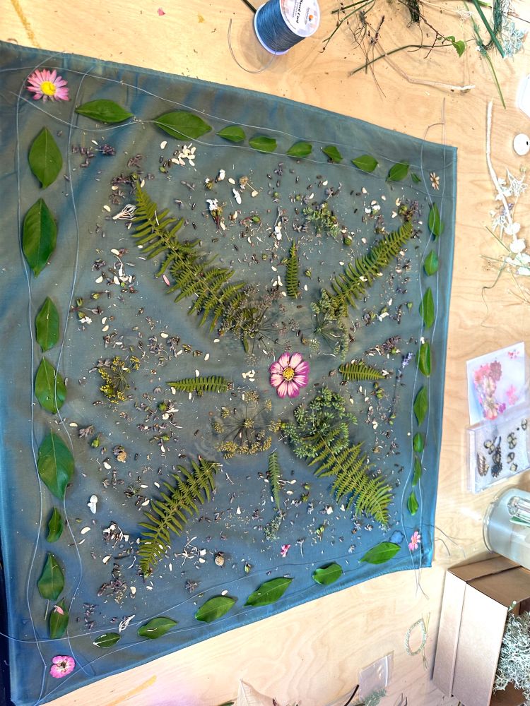 A blue cotton scarf with ferns, leaves, flowers, string, and tiny plant matter arranged on top in an intricate pattern