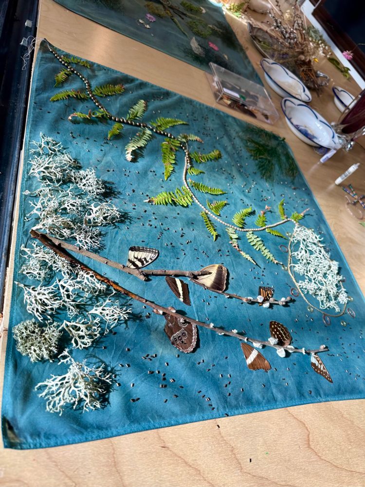 A blue scarf with butterfly wings, willow buds, ferns, lichens, pearls, and beads arranged in a design on top
