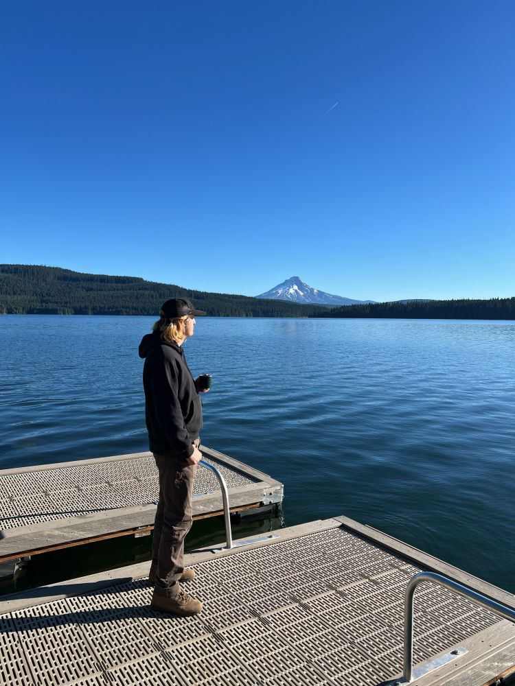 Photograph of me. I am standing on a dock with a cup of coffee overlooking a large lake surrounded by firs. Mt. Hood is visible in the background. 