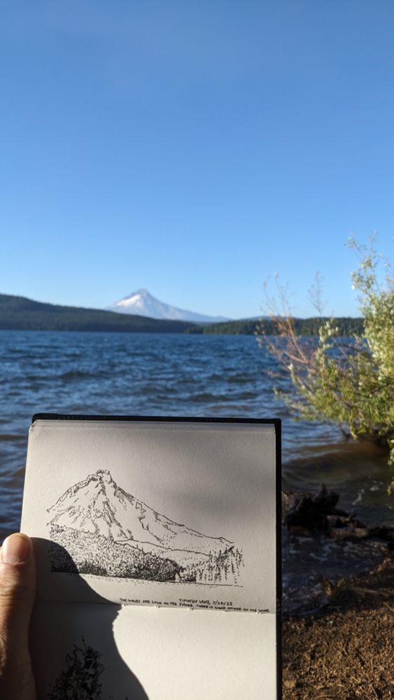 Photograph of an ink sketch of Mt. Hood being held up in front of the actual Mt. Hood, out of focus on the background. 