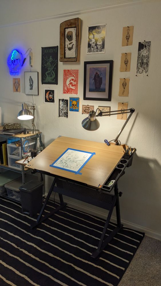 Photo of my workspace with a drawing desk and art supplies. The wall behind it is covered in a variety of skeleton-based art