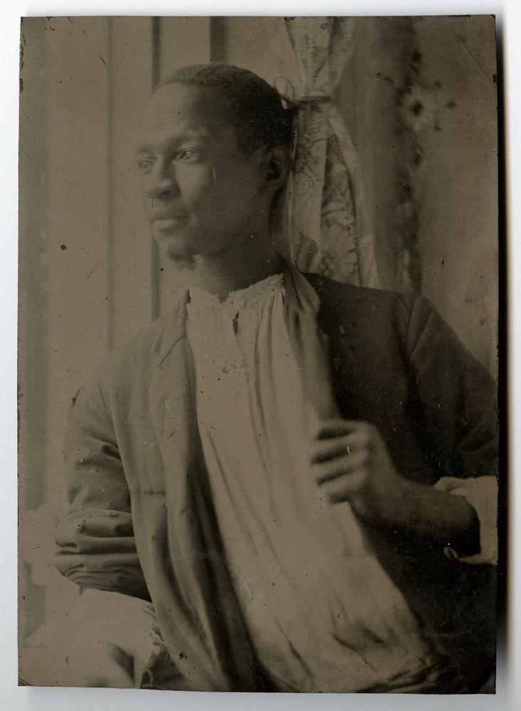 Tintype depicting a half length portrait of a young Black man wearing a dark jacket and a loose-fitting light-colored shirt. He is clean-shaven except for a goatee on his chin, has short hair, and gazes to his right out a window. His left hand is held in front of him in a loose fist. Behind him is a lace curtain.
