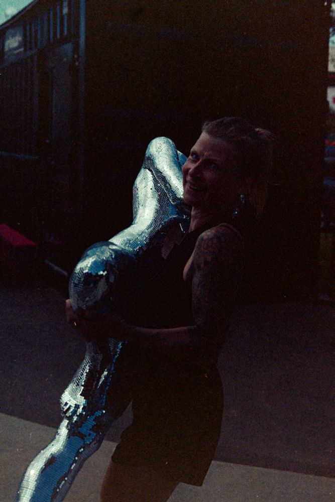 Colour film photo of a woman carrying a mannequin in a standing yoga pose, covered in small mirrors. The photo is somewhat underexposed, so the mannequin is the brightest object in the scene.

Photo by Jacy Eberlein, taken in Edmonton, Alberta on July 1st 2025. Taken with a Nikon F3 with the 50mm f1.4 AFD lens, and Kodak 400.