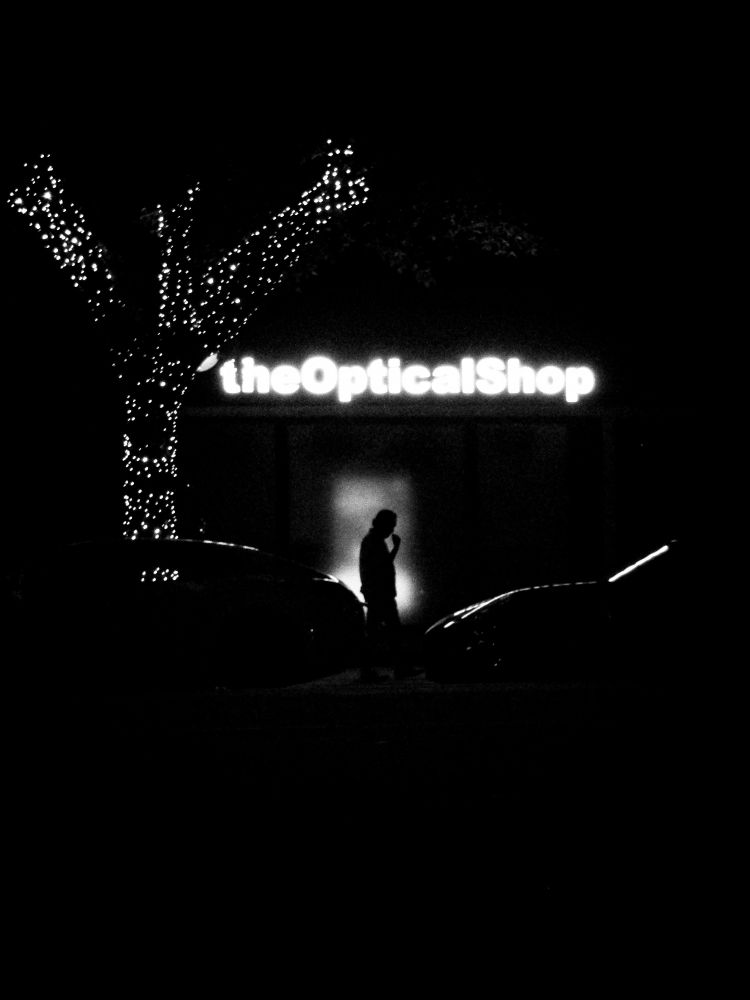 Black and white photo of a person in silhouette against an advertisement being played behind frosted glass. The name of the shop hangs above them, and a tree covered in lights stretches to their left.

Taken in September 2025 by Jacy Eberlein in Edmonton, Alberta. Taken on a Sony DSC-T99.