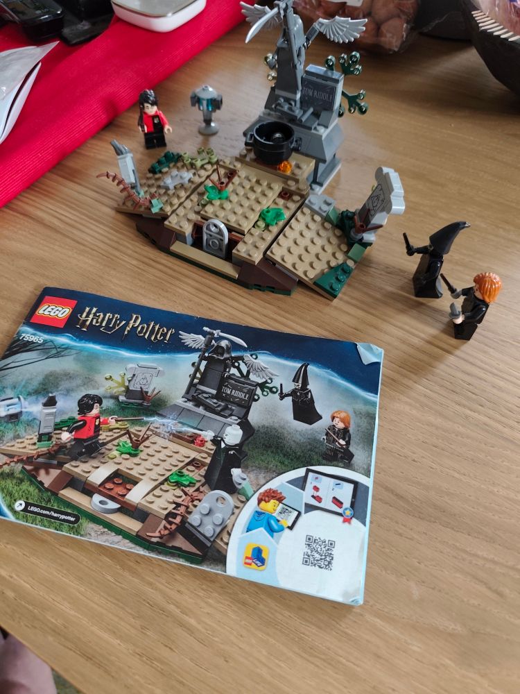 Alt-Text:

Ein LEGO Harry Potter Set ist auf einem Holztisch aufgebaut. Es zeigt eine Szene aus dem Friedhof von Little Hangleton, mit einem Grabstein mit der Aufschrift "Tom Riddle", einer Statue mit Engelsflügeln und mehreren LEGO-Figuren. Zu sehen sind unter anderem Harry Potter, ein Todesser, Voldemort und Cedric Diggory. Im Vordergrund liegt die Bauanleitung des Sets (Setnummer 75965), die das fertige Modell und die enthaltenen Figuren zeigt. Im Hintergrund sind einige Alltagsgegenstände wie eine Schale mit Nüssen und ein roter Tischläufer erkennbar.