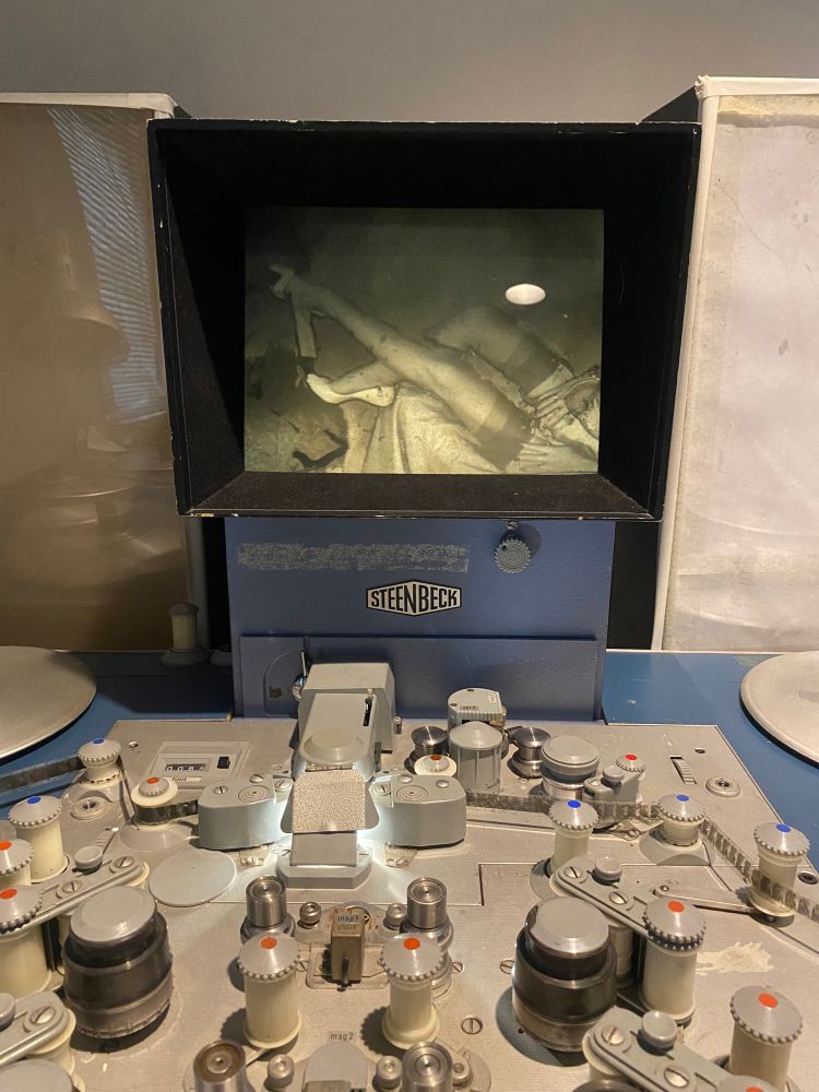 A flatbed steenbeck editor for 16mm film. There are multiple rollers that allow the film to wrap around and pass it through to the viewer, where it is then seen on a screen. On the screen there are a pair of women’s legs with stockings and white high heels.