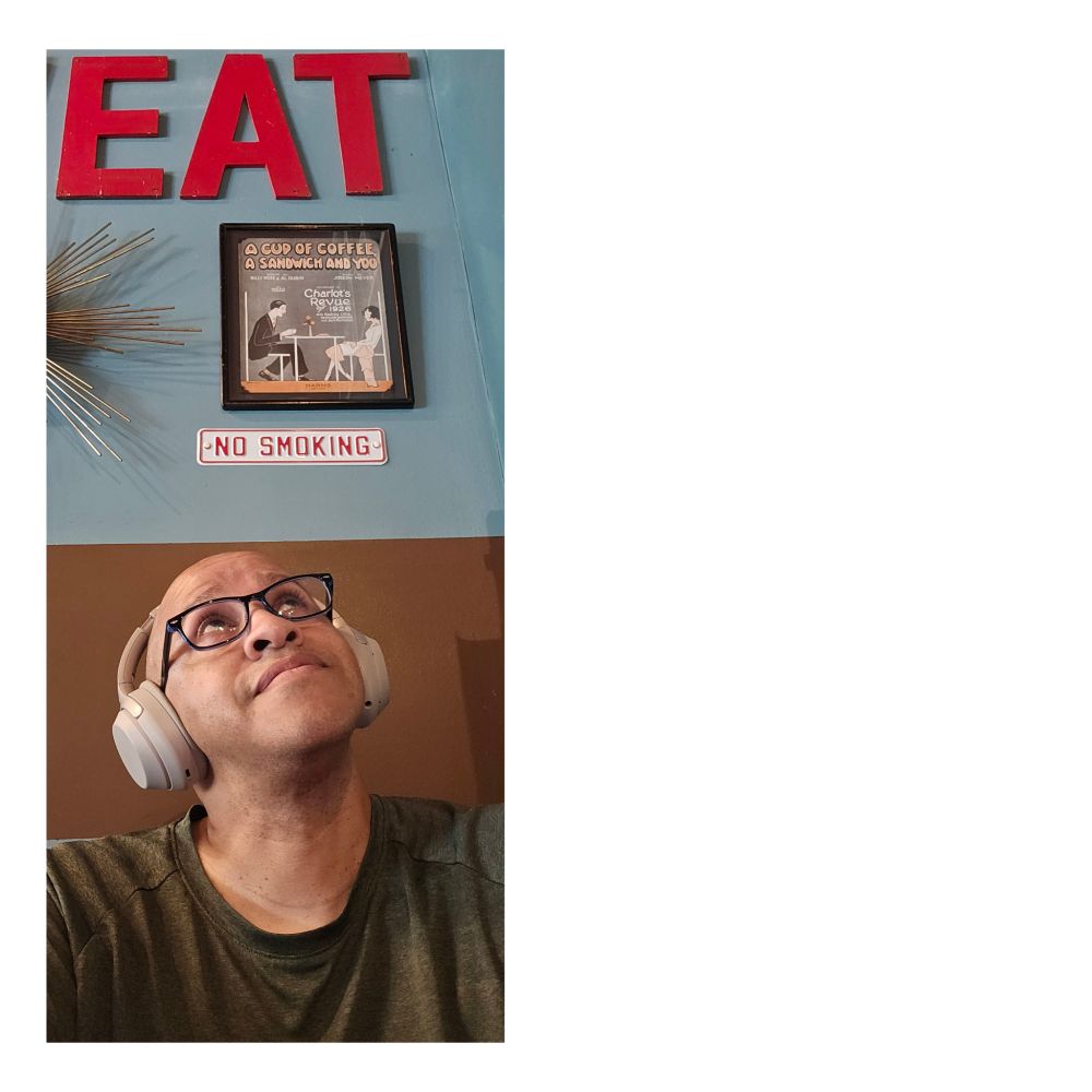 I'm wearing headphones and glasses, looking upward while sitting in a booth at The Mud House coffee shop. The word EAT hangs on the wall behind me 