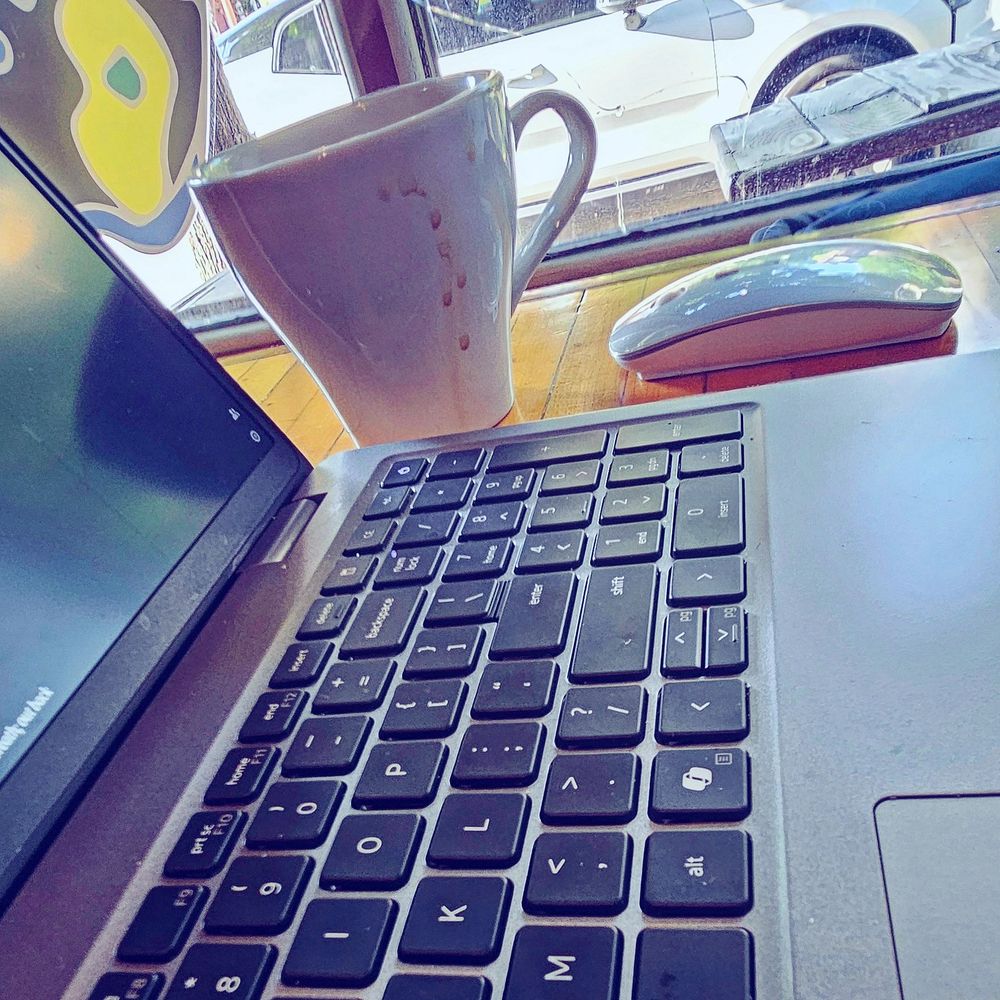 An open laptop sits on a desk in the foreground. Next to it are a coffee cup with dribbles of coffee down its side and computer mouse. In the background is a window with a street view.